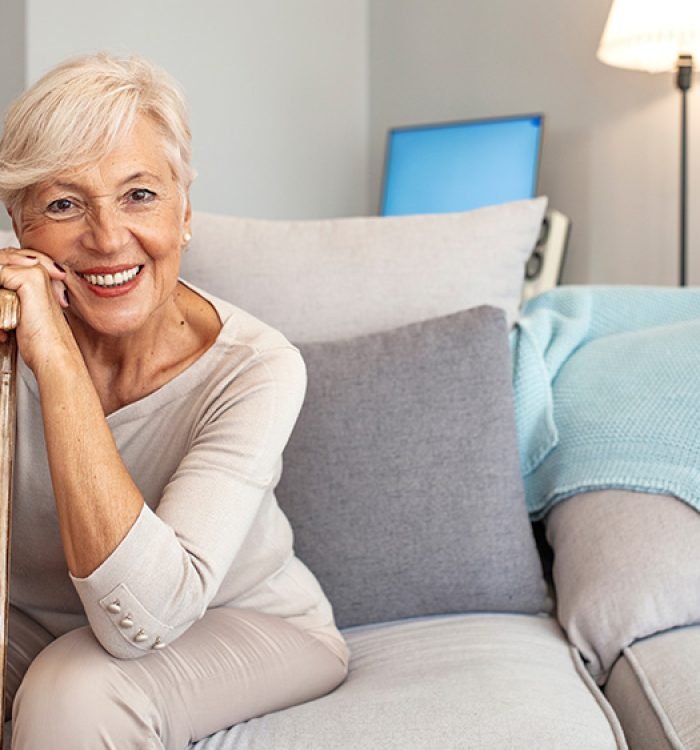 Smiling grandmother sitting on couch. Portrait of a beautiful smiling senior woman with walking cane on light background at home. Old woman sitting with her hands on a cane