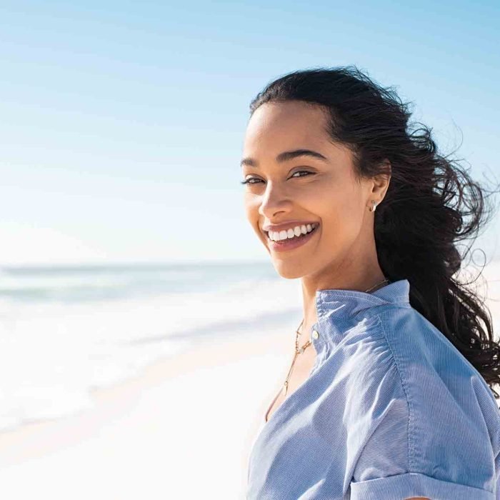 Portrait of young woman at sea looking at camera. Smiling latin hispanic girl standing at the beach with copy space and looking at camera. Happy mixed race girl in casual outfit with wind in her hair.