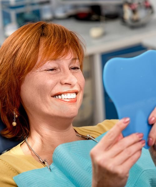 Smiling senior woman with new dental implants sitting in the dental office and looking at the mirror